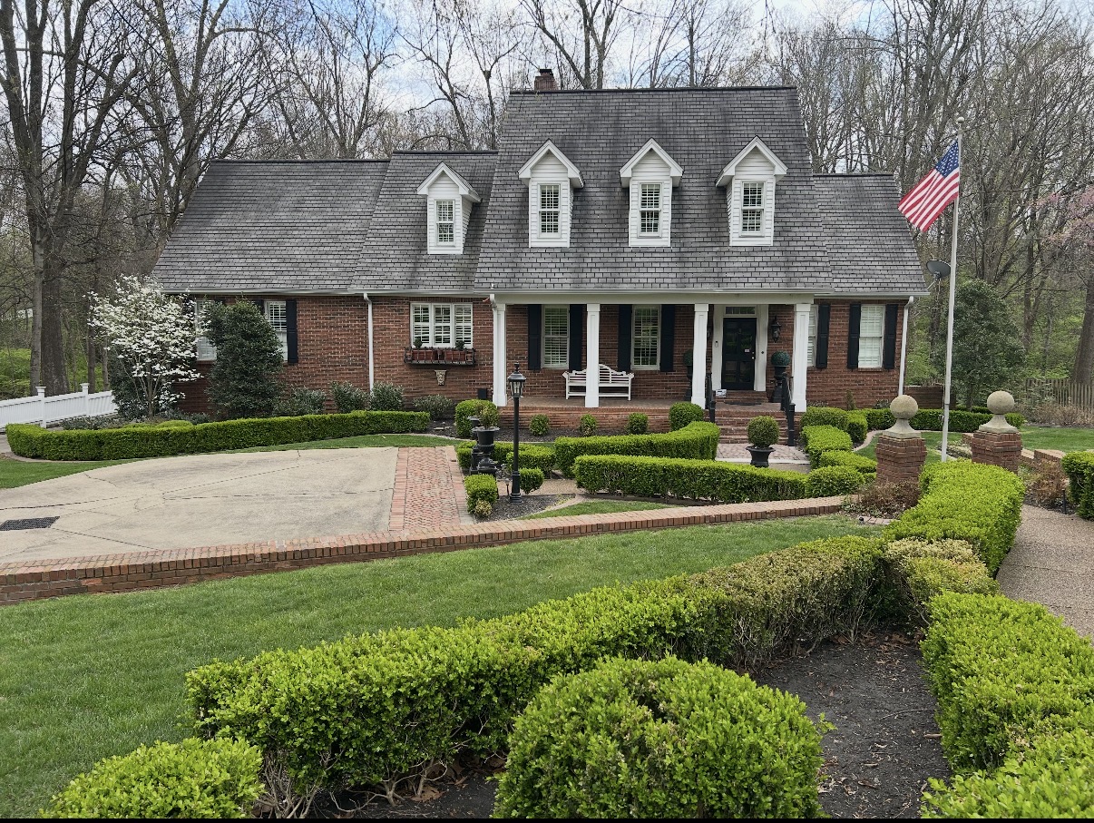 Two-story red brick home that has covered front porch with columns and dormers on second floor. Lovely hardscape and landscaping